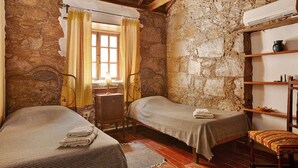A rustic room with two single beds, a wooden chair, a small table, and a stone wall.