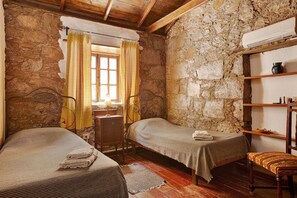 A rustic room with two single beds, a wooden chair, a small table, and a stone wall.
