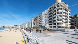 Ein Strandhotel mit mehreren Balkonen, im Hintergrund ein Strand und eine Promenade mit Spaziergängern.