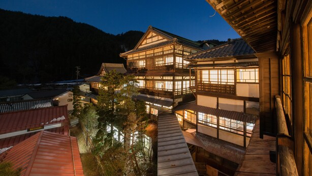 Ein traditionelles japanisches Gebäude mit mehreren Ebenen und großen Fenstern, umgeben von Bäumen und einem Bergpanorama.