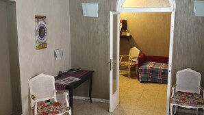 A room with a door leading to another room, a small table with a newspaper, two white wicker chairs, and a red sofa with a plaid blanket.