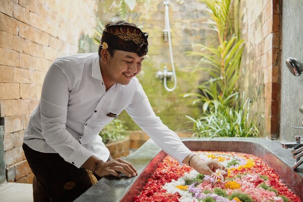 Una persona con camisa blanca y pantalones negros inclinada sobre una bañera grande llena de pétalos de flores de colores.