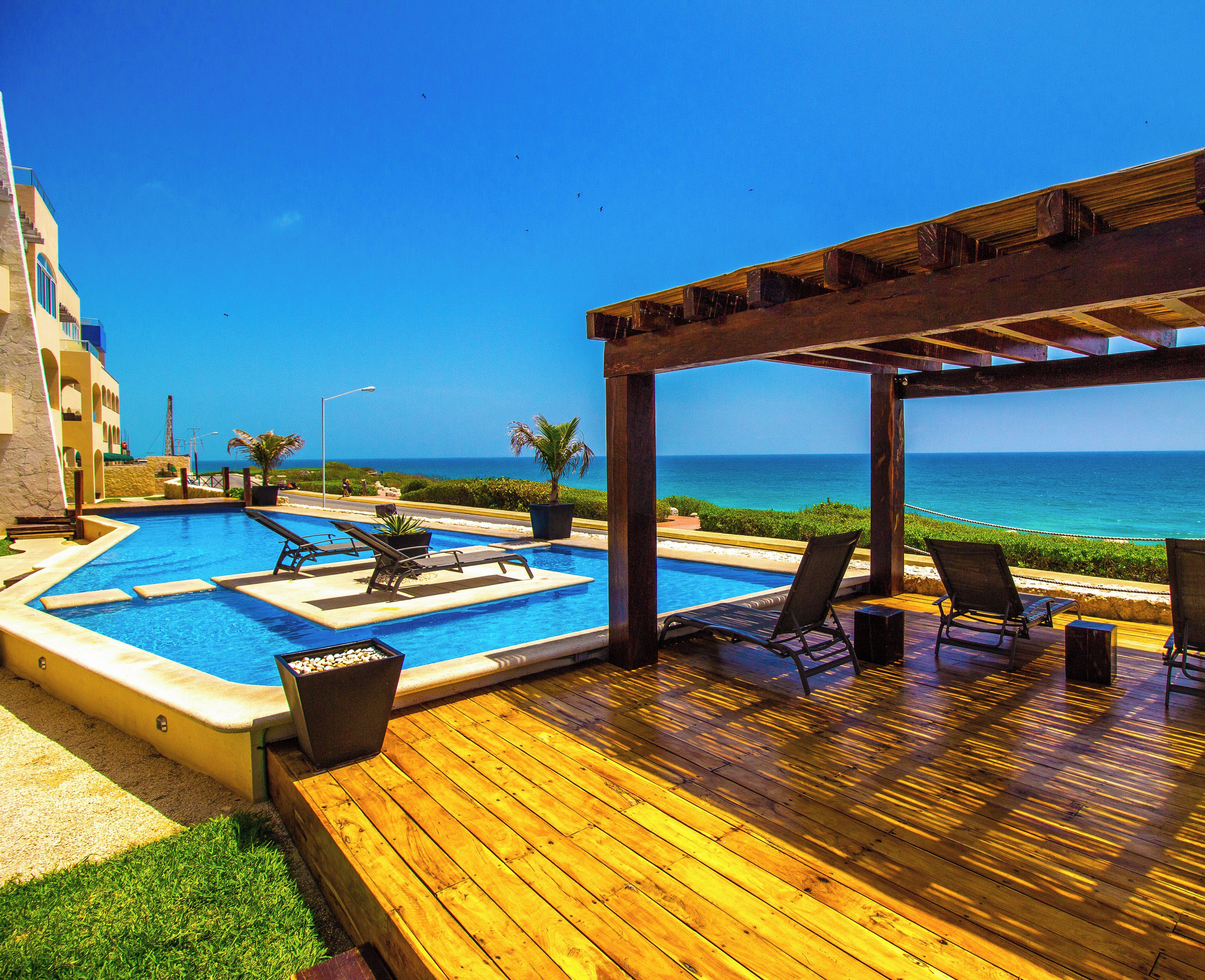 A wooden deck with a pool, lounge chairs, and a view of the ocean.