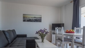Un salon avec un canapé noir, une table blanche et un téléviseur. Il y a un tableau au mur et une fenêtre avec des rideaux.