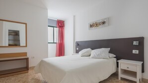Une chambre à coucher comprenant un lit, des tables de chevet, un banc, une fenêtre avec un rideau rouge et un luminaire fixé au mur.
