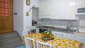 Une cuisine avec des meubles blancs, un four à micro-ondes et un évier. On y trouve une table avec un panier de fruits et une nappe à motifs de tournesols.