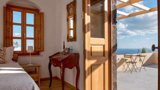 A bedroom with a large bed, a wooden desk, a mirror, and a view of the sea through an open door.