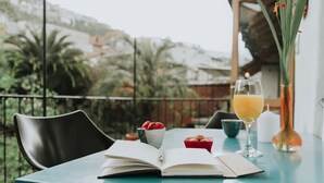 A table with an open book, a glass of orange juice, a bowl of strawberries, and a small red box of cookies on a balcony with a view of greenery and buildings.