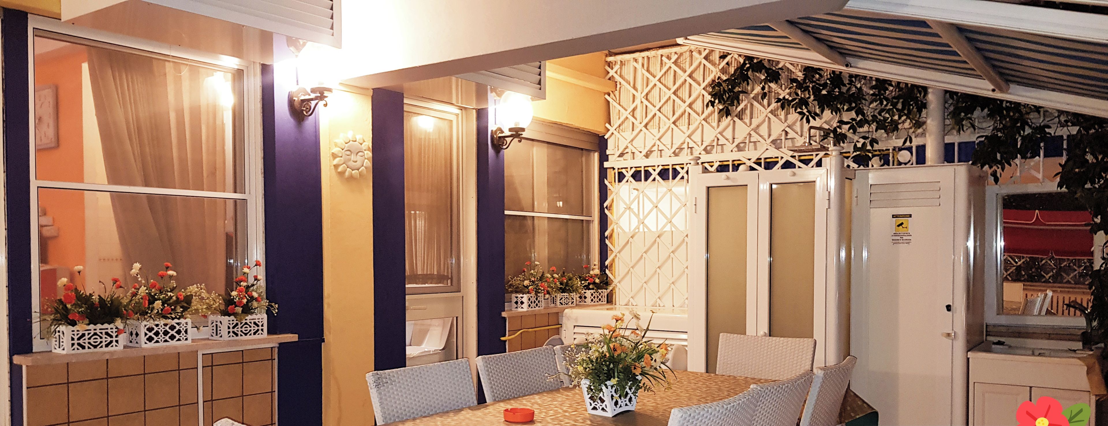 Ein Essbereich mit einem Tisch, der für sechs Personen gedeckt ist, umgeben von weißen Stühlen. Auf den Fensterbänken stehen Blumenarrangements und eine Wand ist mit einem Gittermuster verziert.