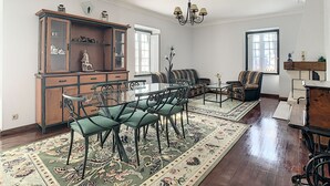A spacious living room with a wooden cabinet, green upholstered chairs, a glass coffee table, and a patterned rug.