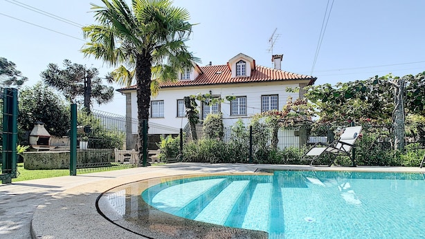 A swimming pool with a curved edge, a white house with a red roof in the background, and a palm tree.