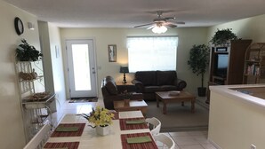 Uma sala de jantar com uma mesa branca, jogos americanos vermelhos e verdes, um vaso de flores amarelas e cadeiras brancas.