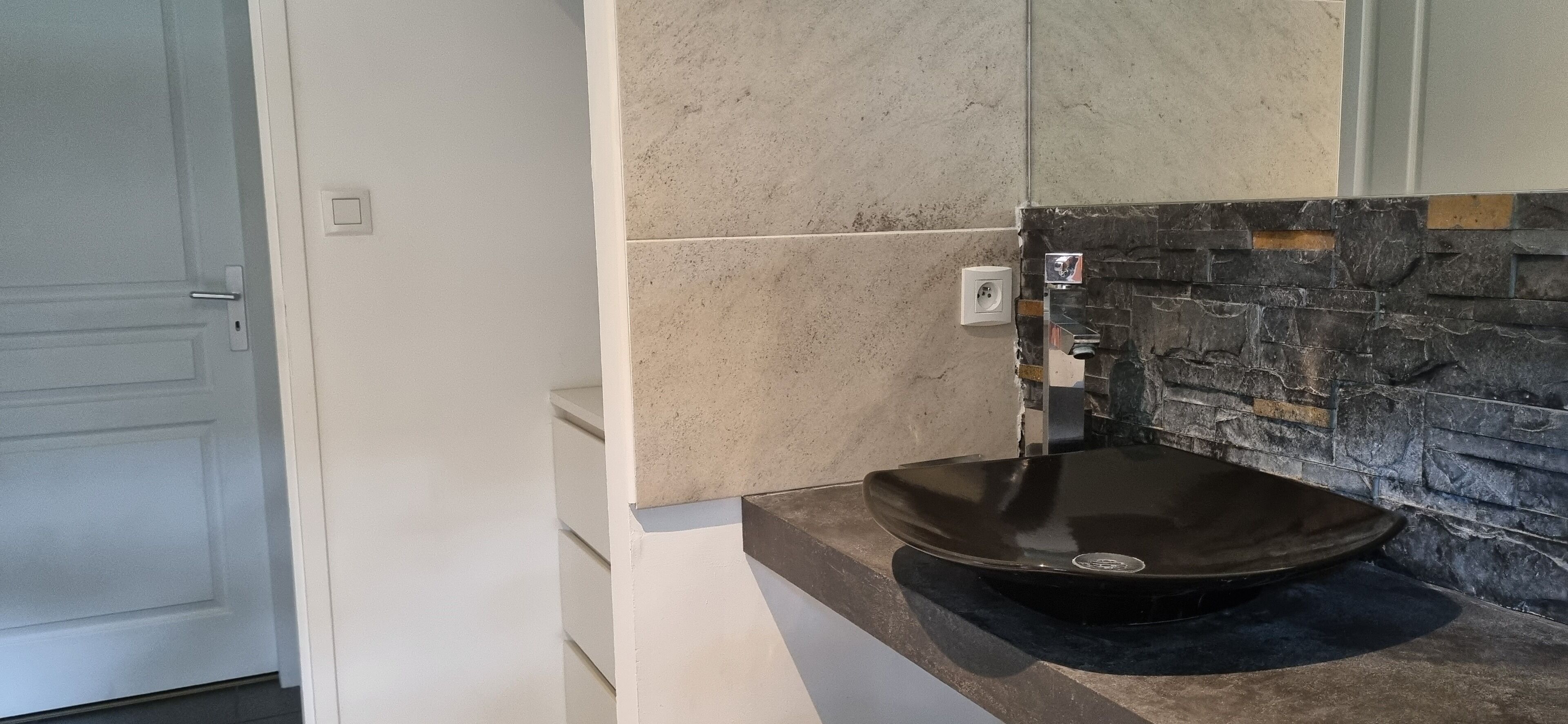 Une salle de bain avec un lavabo sur colonne noir, une porte blanche et un mur recouvert de pierres apparentes.