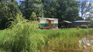 Een rustieke blokhut met een veranda en een klein tuinhuisje, omgeven door weelderige begroeiing en een vijver.