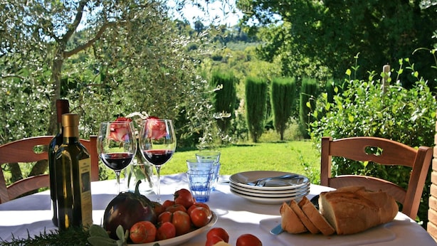 Ein Tisch im Freien mit einer Weinflasche, Gläsern, einer Tomatenschale, einem Brotlaib und einer kleinen Käseschale.