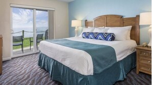 Een hotelkamer met een groot bed, een houten hoofdbord, een nachtkastje, een lamp, en uitzicht door een schuifdeur op een buitenruimte met stoelen.