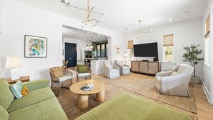 Ein geräumiges Wohnzimmer mit grünen Sofas, einem Couchtisch und einem Fernseher. Im Hintergrund befindet sich eine Küche mit weißen Schränken und ein Essbereich mit Tisch und Stühlen.
