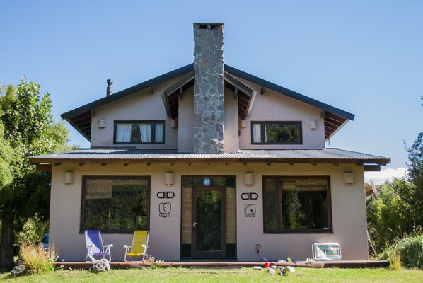 A two-story house with a stone chimney, large windows, and a covered porch with two lawn chairs.