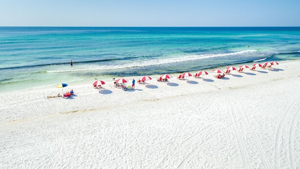 Een strand met wit zand en helderblauw water. Er staan rode parasols en enkele mensen zijn zichtbaar.