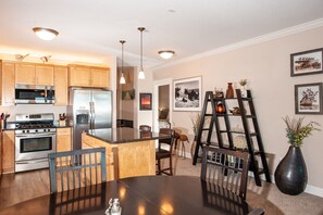 A modern kitchen with stainless steel appliances, wooden cabinets, and a central island. There is a dining area with a dark table and chairs.