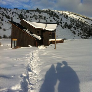 Una cabaña de madera con techo nevado, entrada visible y un pequeño porche.