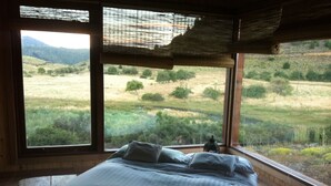 A bedroom with a large bed, a window with a view of a grassy field and trees, and a wooden ceiling. - Your Home In The Andes (San Martin de los Andes)