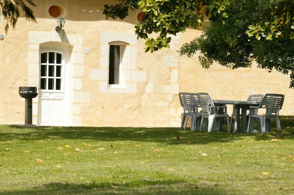 Un bâtiment avec une façade en pierre, une porte blanche et une fenêtre. On y trouve des chaises en plastique et une table à l’extérieur.