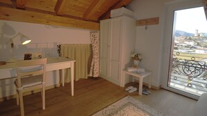 Une pièce avec des poutres en bois au plafond, un bureau avec une lampe, une chaise, une armoire, une petite table avec un vase et un balcon offrant une vue sur la ville et les montagnes.