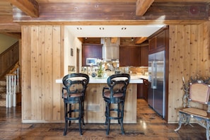 Una cucina con rivestimenti in legno, due sgabelli da bar, un frigorifero in acciaio inossidabile e una sedia decorativa.