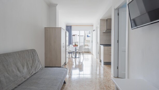 Un ambiente moderno e luminoso con un divano grigio, una TV a schermo piatto fissata al muro e una zona cucina visibile attraverso una porta aperta.
