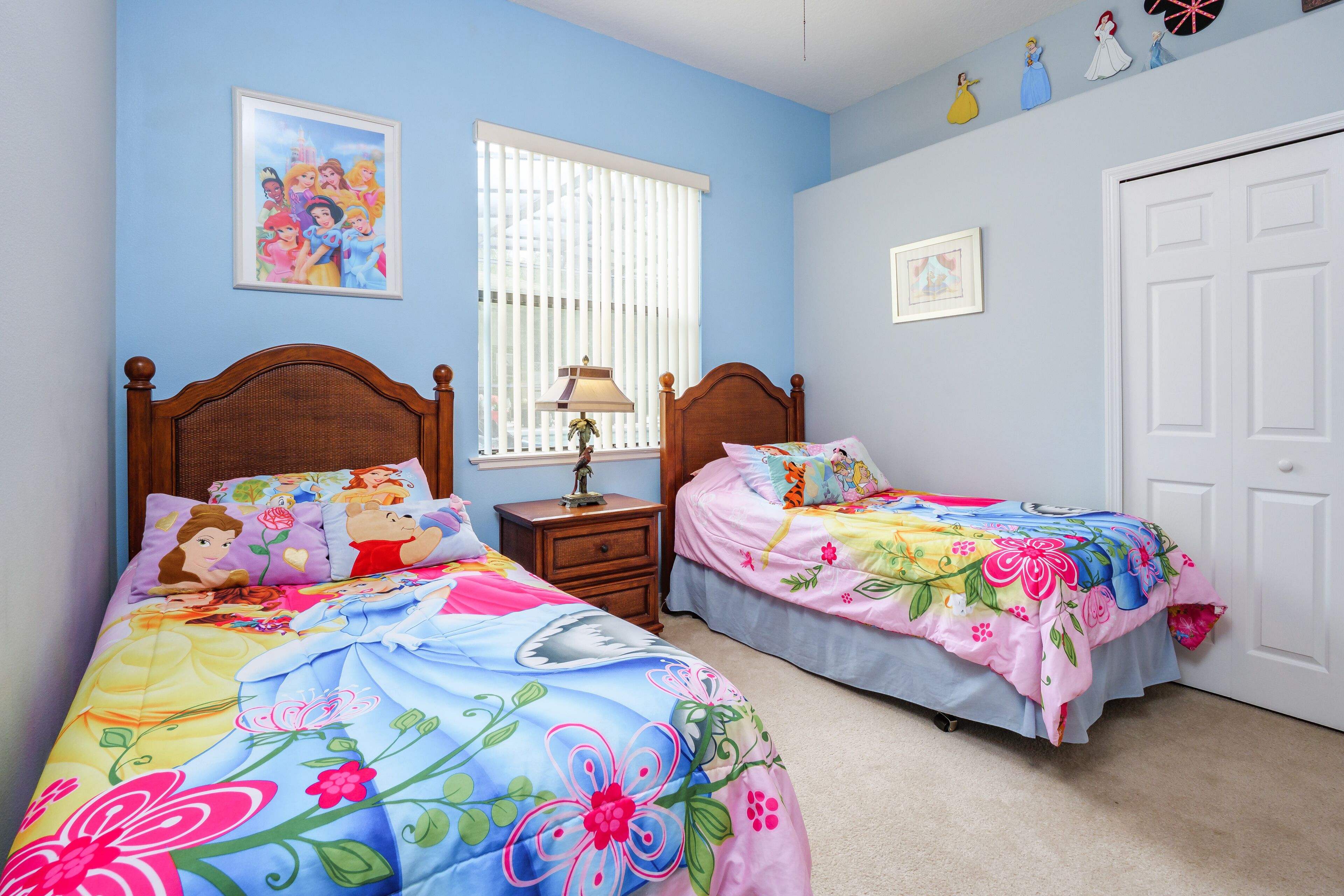 A bedroom with two beds, each with a Disney-themed bedspread, a nightstand with a lamp, and a window with blinds.