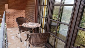 Een balkon met twee rieten stoelen en een kleine ronde tafel, een bakstenen muur en grote ramen met uitzicht op bomen.