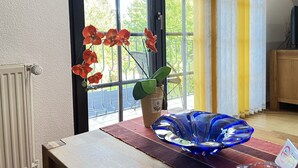 Een kamer met een raam, een radiator, een tafel met een vaas rode bloemen en een blauw glazen object.