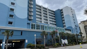 Ein mehrstöckiges Hotel mit blauer Fassade, einem überdachten Parkplatz und Palmen.