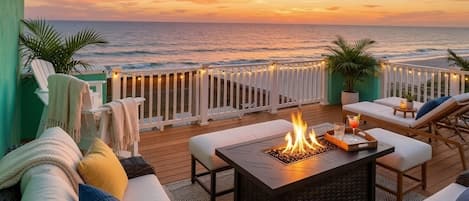 A deck with outdoor seating, a fire pit, and a view of the ocean at sunset.