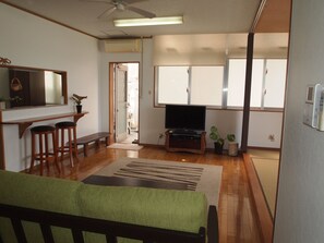 A living room with a television, wooden flooring, a bench, and a green sofa. - Rental Villa Lana Waioli (Tomigusuku)
