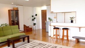 A living room with a green sofa, a wooden coffee table, and a striped rug. There is a kitchen area with a white countertop and two stools. - Rental Villa Lana Waioli (Tomigusuku)