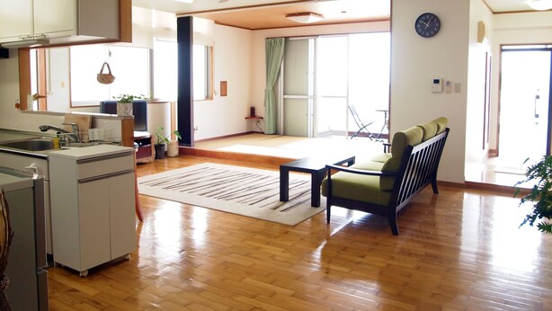 A spacious living area with a kitchen, wooden flooring, a ceiling fan, and a large sliding glass door leading to a balcony. - Rental Villa Lana Waioli (Tomigusuku)
