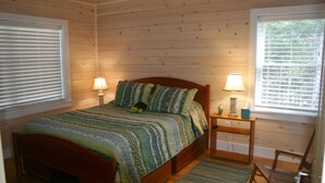 A wooden-paneled bedroom with a bed, two bedside lamps, a nightstand, a chair, and a window with blinds. - Tranquil oceanfront cottage on Wohoa Bay (Addison)