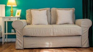 Un canapé beige agrémenté de deux coussins, adossé à un mur de couleur turquoise, une petite table de chevet blanche avec une lampe et un tableau encadré.