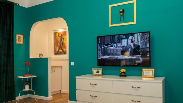 Une pièce aux murs de couleur turquoise, avec une télévision, une commode blanche, une petite table sur laquelle est posé un vase, et une porte qui donne sur une autre pièce.