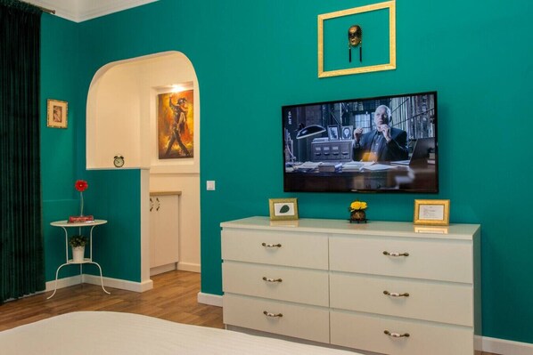 Ein türkisfarbenes Zimmer mit einem Fernseher, einer weißen Kommode, einem kleinen Tisch mit einer Vase und einer Tür zu einem anderen Raum.