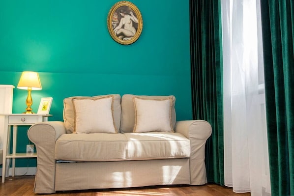 Ein Zimmer mit einer türkisfarbenen Wand, einem beigen Sofa mit zwei Kissen, einem kleinen weißen Beistelltisch mit einer Lampe und einem gerahmten Bild über dem Sofa.