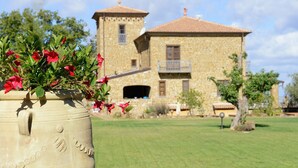 Un grand pot en céramique orné de fleurs rouges, posé sur une surface en pierre. En arrière-plan, on aperçoit un bâtiment en pierre doté d’un balcon, avec un ciel dégagé.