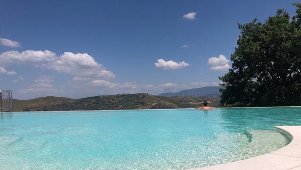 Une piscine d’un bleu éclatant où une personne nage, entourée d’une zone pavée et offrant une vue sur des collines et des arbres.
