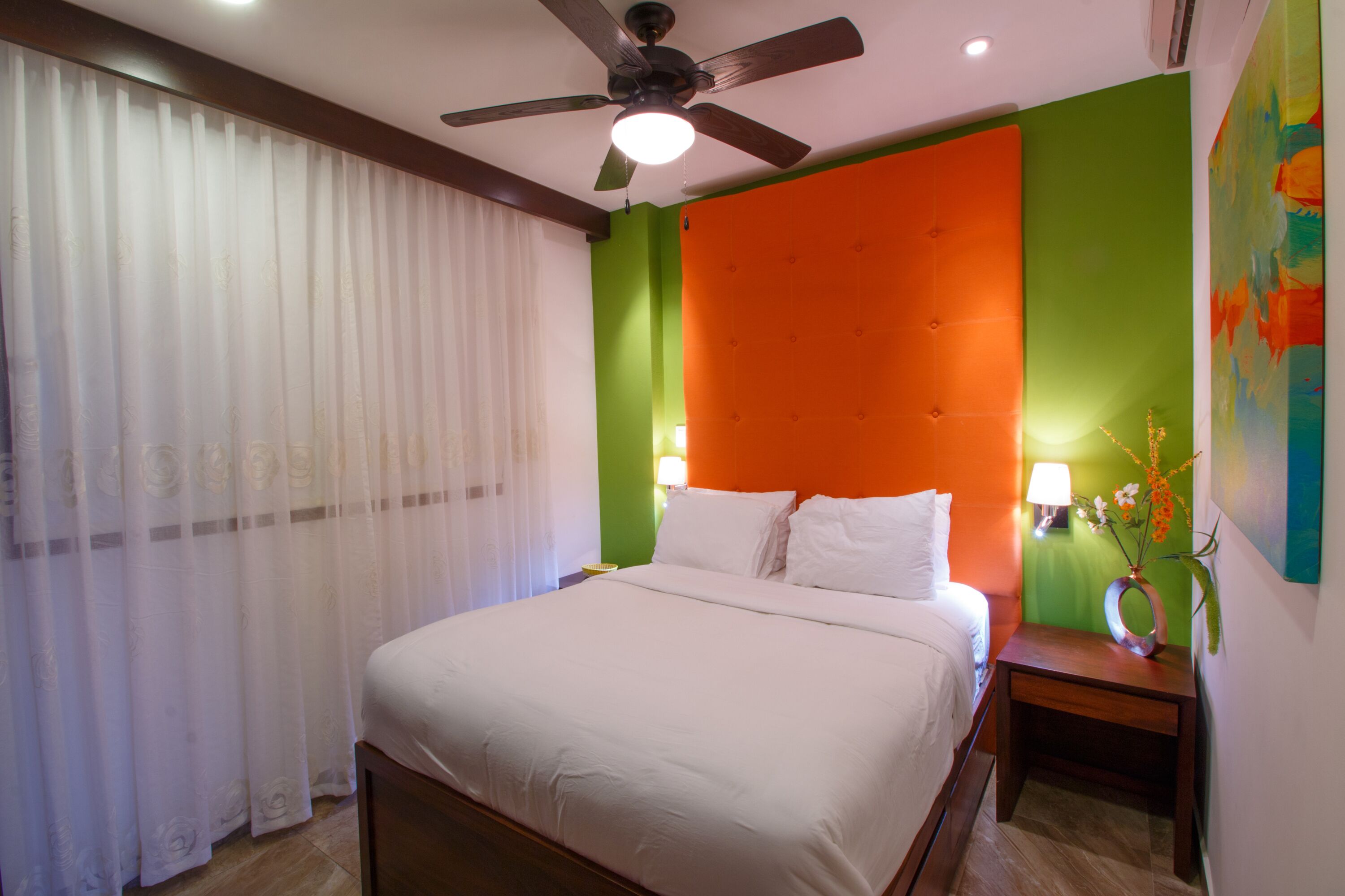 Habitación de hotel con cama, ventilador de techo, mesita de noche con lámpara y un cuadro colorido en la pared.