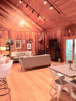 A cozy cabin interior with a wooden ceiling, a sofa, a dining table, and a TV. - Oxford Retreat - 4B/4B Newly Renovated Cabin 15 Minutes From Downtown Oxford! (Batesville)