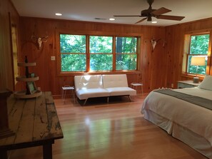 A wooden cabin room with a white sofa, a bed, a small table, and a ceiling fan. - Oxford Retreat - 4B/4B Newly Renovated Cabin 15 Minutes From Downtown Oxford! (Batesville)