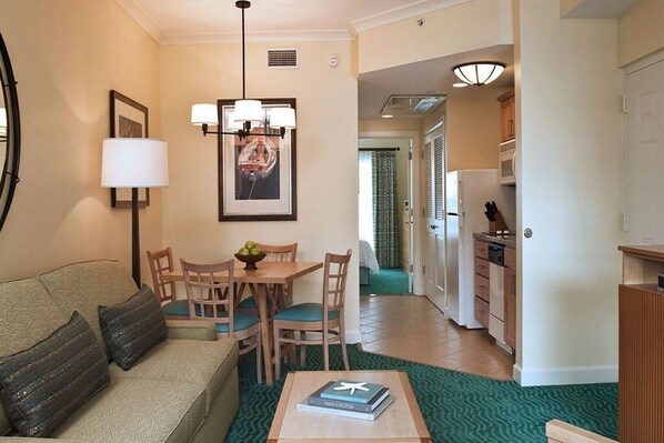 A living room with a sofa, a dining table with chairs, a coffee table with a book, a kitchen area with cabinets and a refrigerator, and a hallway leading to another room.