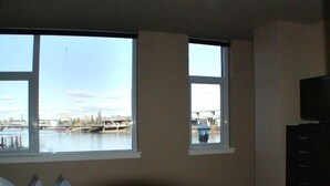 Slaapkamer met een groot raam met uitzicht op een waterlichaam en een brug, een bed met kussens, en een televisie op een standaard.